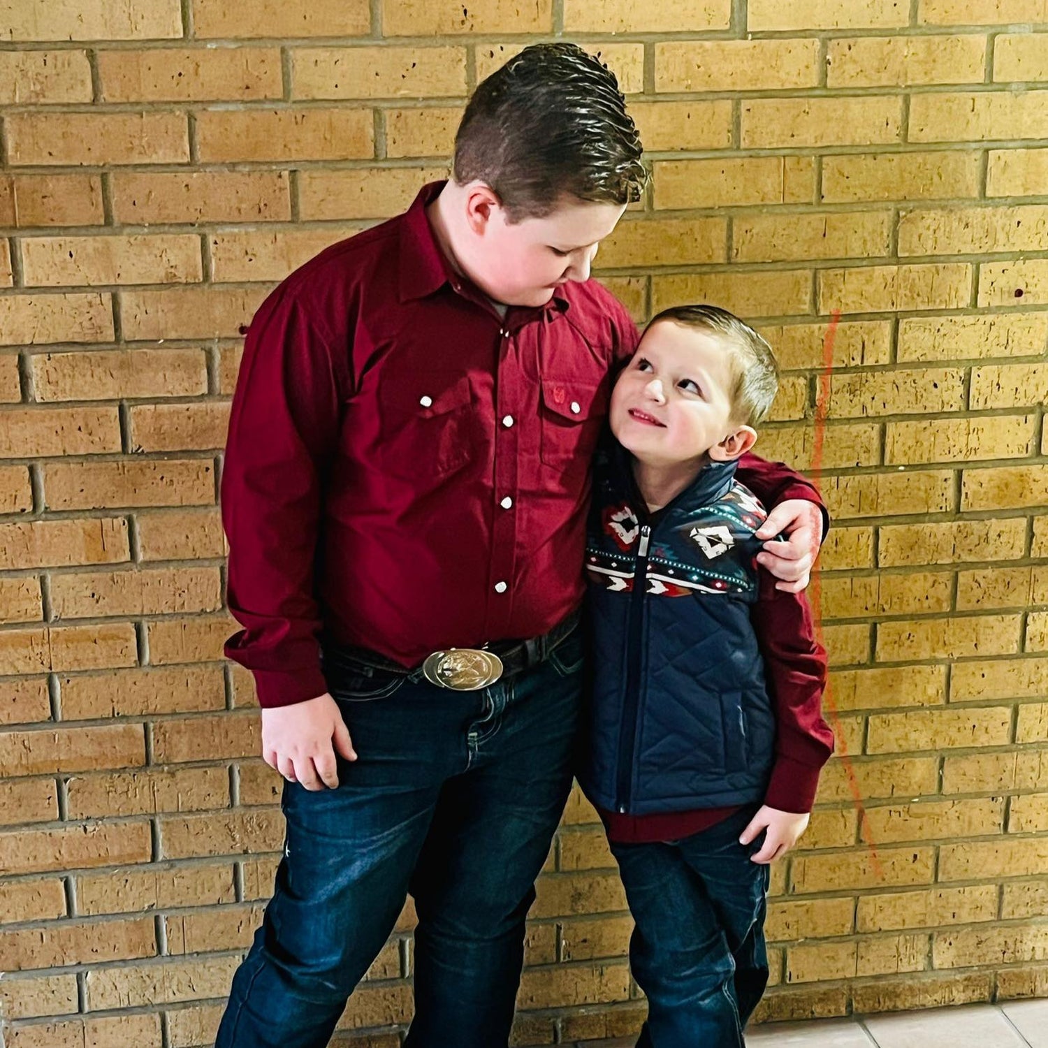 Male child in bootcut boys denim and another male child in western bootcut denim and long sleeve pearl snap button down with western style vest.  Both in cowboy boots