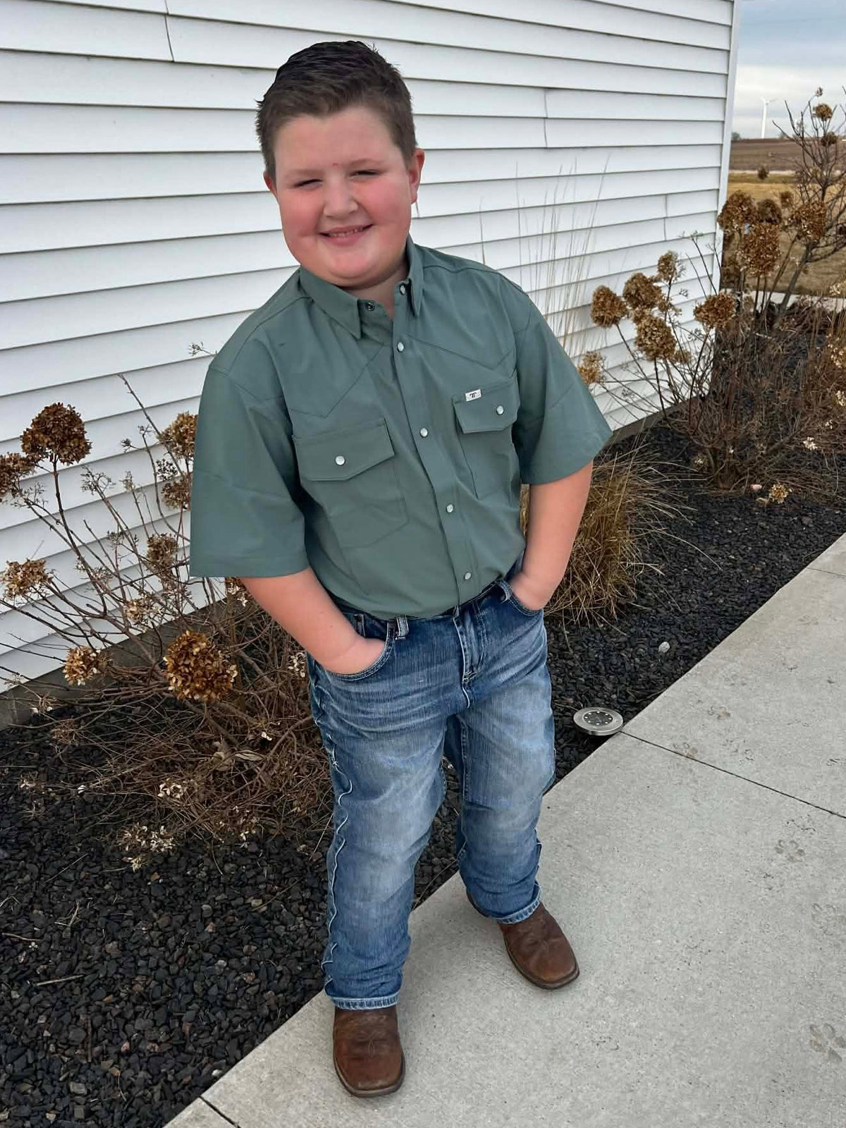 A young boy wearing a green polo with denim and cowboy boots that also comes in men size for church, showing livestock, or everyday